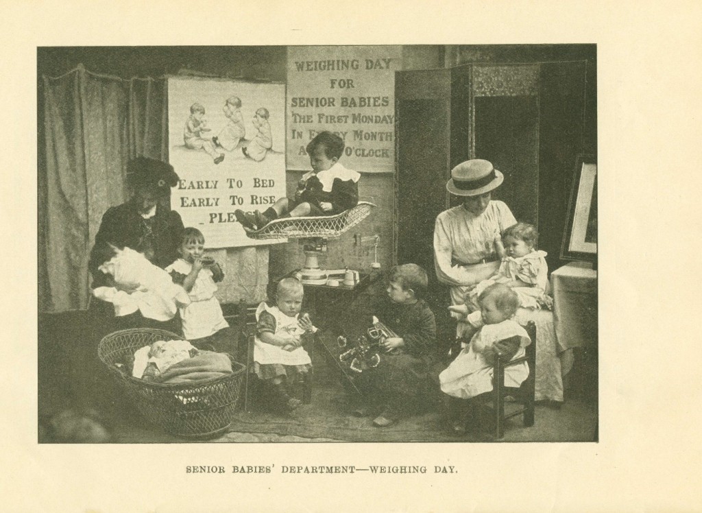 Image of two women with babies and small children. One small boy is sat in a basket on top of weighing scales. 